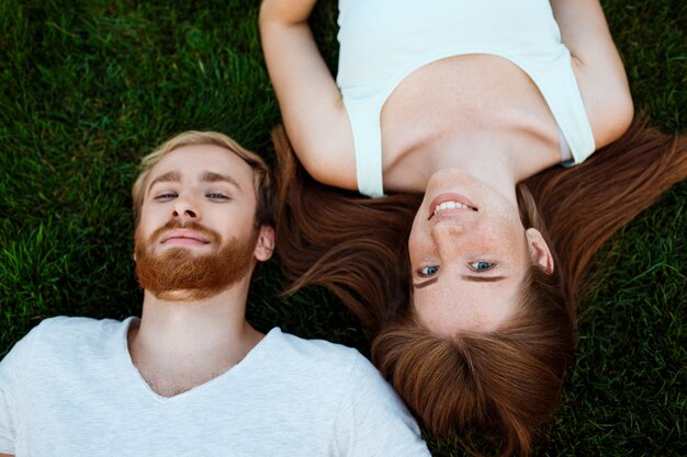 Joven pareja hermosa sonriendo, tumbado en la hierba en el parque. Disparo desde arriba.