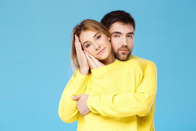 Joven pareja hermosa en un suéter amarillo abrazando sonriendo sobre pared azul