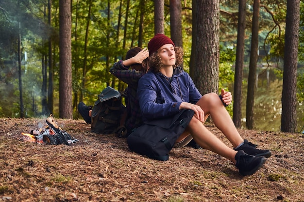 Joven pareja hipster haciendo senderismo en el bosque y sentándose espalda con espalda mientras se calienta cerca de una fogata en el campamento. Concepto de viaje, turismo y caminata.