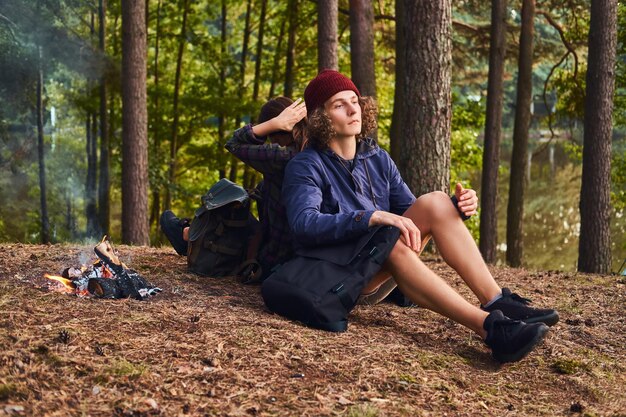 Joven pareja hipster haciendo senderismo en el bosque y sentándose espalda con espalda mientras se calienta cerca de una fogata en el campamento. Concepto de viaje, turismo y caminata.