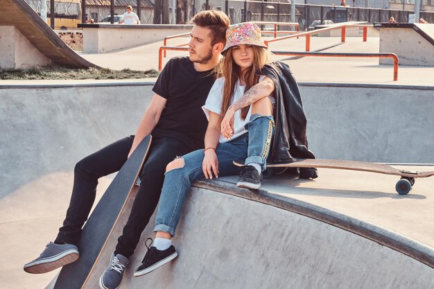 Joven pareja hipster vestida de moda relajándose junto con patinetas en un parque de patinaje en un día soleado.