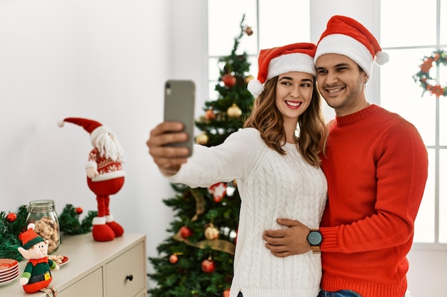 Joven pareja hispana sonriendo feliz usando sombrero de navidad haciendo selfie por el teléfono inteligente en casa.