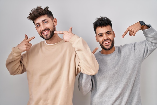 Joven pareja homosexual de pie sobre fondo blanco sonriendo alegre mostrando y señalando con los dedos los dientes y la boca concepto de salud dental