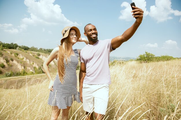 Joven pareja internacional multiétnica al aire libre en la pradera en un día soleado de verano. Hombre afroamericano y mujer caucásica haciendo picnic juntos. Concepto de relación, verano. Haciendo selfie.