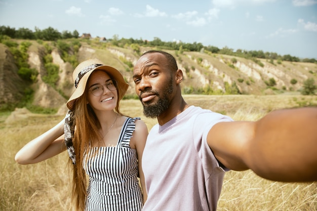 Joven pareja internacional multiétnica al aire libre en la pradera en un día soleado de verano. Hombre afroamericano y mujer caucásica haciendo picnic juntos. Concepto de relación, verano. Haciendo selfie.