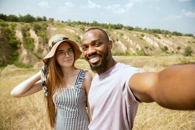 Joven pareja internacional multiétnica al aire libre en la pradera en un día soleado de verano. Hombre afroamericano y mujer caucásica haciendo picnic juntos. Concepto de relación, verano. Haciendo selfie.