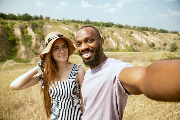 Joven pareja internacional multiétnica al aire libre en la pradera en un día soleado de verano. Hombre afroamericano y mujer caucásica haciendo picnic juntos. Concepto de relación, verano. Haciendo selfie.