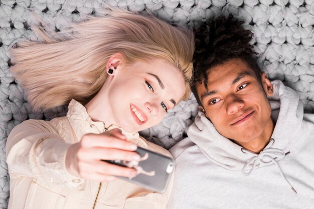 Joven pareja interracial tumbado en la alfombra tomando selfie en teléfono móvil