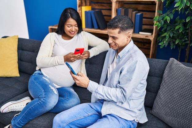 Joven pareja latina esperando un bebé usando un teléfono inteligente sentado en un sofá en casa