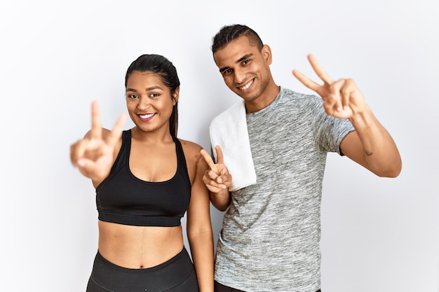 Foto gratuita joven pareja latina con ropa deportiva de pie sobre un fondo aislado sonriendo mirando a la cámara mostrando los dedos haciendo el signo de la victoria número dos