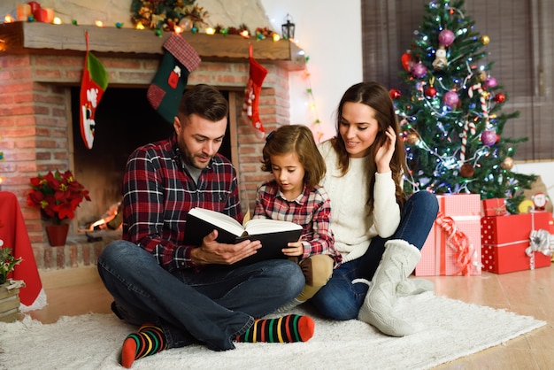 Joven pareja leyendo un libro con una niña en su sala de estar decoradas para la navidad