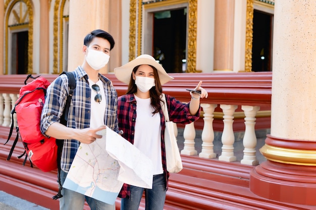 Foto gratuita joven pareja de mochileros asiáticos en un hermoso templo durante las vacaciones en tailandia, una mujer bonita usa un sombrero señalando a donde quieren ir, sostienen un mapa de papel y un teléfono inteligente para verificar la dirección