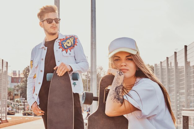 Una joven pareja de moda se está relajando en la calle soleada con sus longboards. Ellos usan mezclilla.