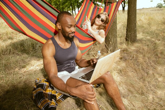 Joven pareja romántica internacional multiétnica al aire libre en la pradera en un día soleado de verano. Hombre afroamericano y mujer caucásica haciendo picnic juntos. Concepto de relación, verano.