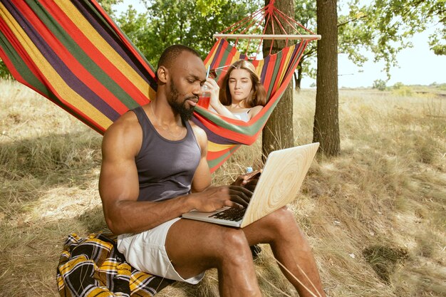 Joven pareja romántica internacional multiétnica al aire libre en la pradera en un día soleado de verano. Hombre afroamericano y mujer caucásica haciendo picnic juntos. Concepto de relación, verano.