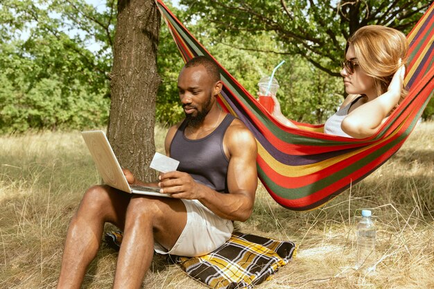 Joven pareja romántica internacional multiétnica al aire libre en la pradera en un día soleado de verano. Hombre afroamericano y mujer caucásica haciendo picnic juntos. Concepto de relación, verano.