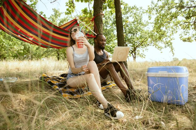 Joven pareja romántica internacional multiétnica al aire libre en la pradera en un día soleado de verano. Hombre afroamericano y mujer caucásica haciendo picnic juntos. Concepto de relación, verano.