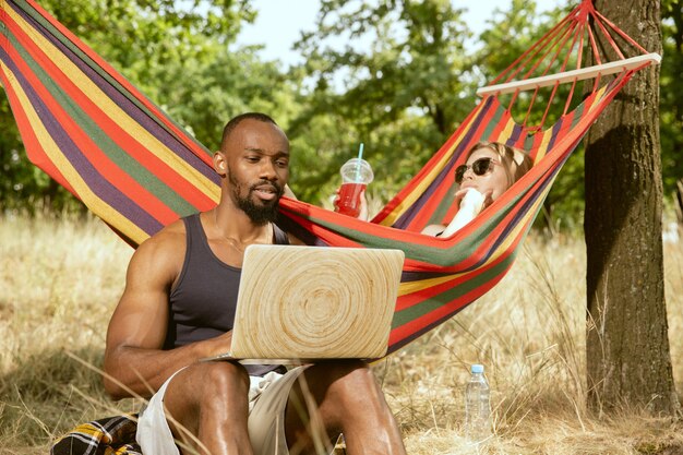 Joven pareja romántica internacional multiétnica al aire libre en la pradera en un día soleado de verano. Hombre afroamericano y mujer caucásica haciendo picnic juntos. Concepto de relación, verano.