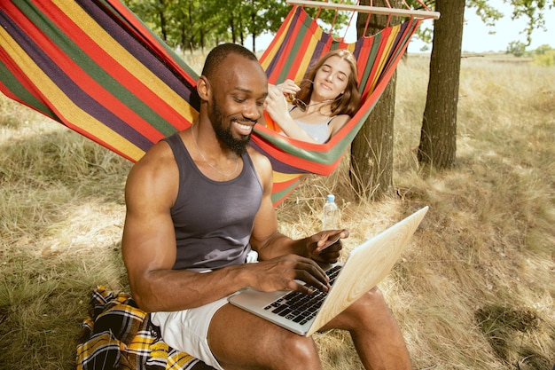 Joven pareja romántica internacional multiétnica al aire libre en la pradera en un día soleado de verano. Hombre afroamericano y mujer caucásica haciendo picnic juntos. Concepto de relación, verano.