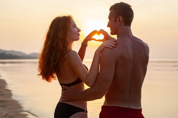 Joven pareja romántica sexy enamorada feliz en la playa de verano juntos divirtiéndose vistiendo trajes de baño mostrando signo de corazón en sundet