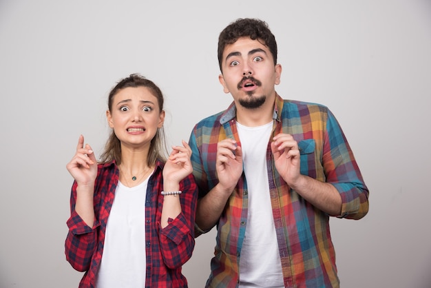 Una joven pareja sobre fondo gris con los dedos cruzados y deseando lo mejor.