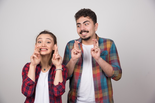 Una joven pareja sobre fondo gris con los dedos cruzados y deseando lo mejor.