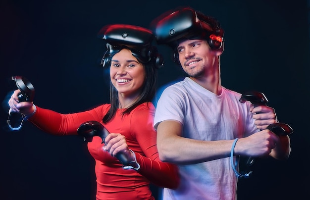 Joven pareja sonriente divirtiéndose juntos, jugando con auriculares y controladores VR. Aislado sobre fondo oscuro. Foto con efecto de luz.