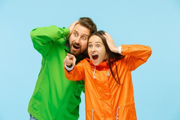 La joven pareja sorprendida apuntando a la izquierda y posando en el estudio en chaquetas de otoño aisladas en azul. Emociones humanas negativas. Concepto de clima frío. Conceptos de moda femenina y masculina