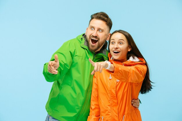 La joven pareja sorprendida apuntando a la izquierda y posando en el estudio en chaquetas de otoño aisladas en azul.