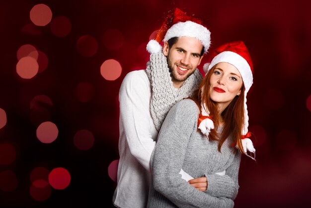 Joven pareja vestida con ropa de invierno y sombrero de santa en un fondo rojo con el bokeh