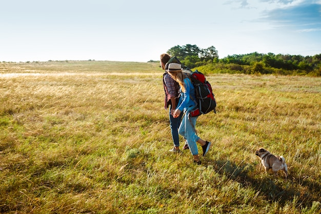 Joven pareja de viajeros caminando en campo con perro pug
