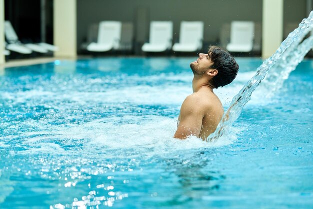 Joven pasando un día en el spa y relajándose bajo un chorro de agua en la piscina