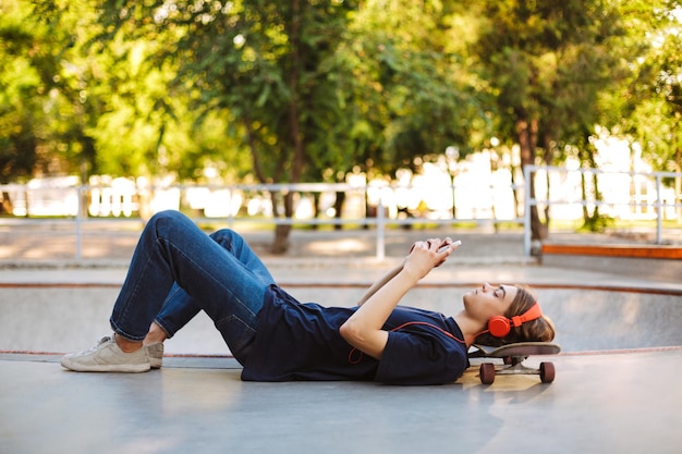Un joven patinador con auriculares naranjas acostado en una patineta cuidadosamente usando un celular con un moderno parque de patinaje en el fondo