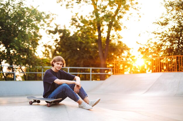 Joven patinador en camiseta negra y jeans mirando soñadoramente en cámara pasando tiempo con patineta en el moderno parque de patinaje con puesta de sol en el fondo