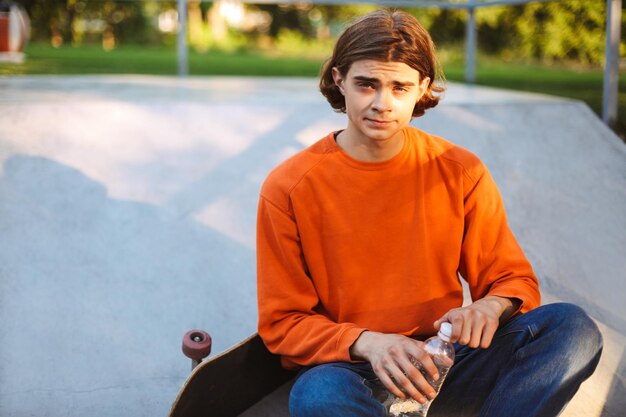 Joven patinador con jersey naranja sosteniendo una botella de agua en las manos mientras mira soñadoramente a la cámara con patineta en el parque de patinaje