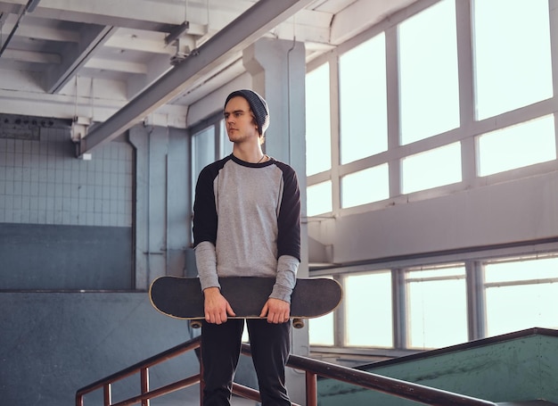 Un joven patinador parado junto a un riel de molienda en un parque de patinaje en el interior, sosteniendo su tabla y mirando hacia otro lado.