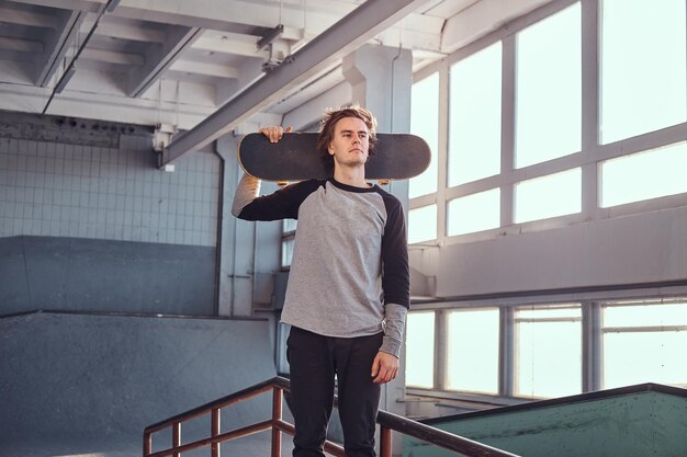 Un joven patinador parado junto a un riel de molienda en un parque de patinaje en el interior, sosteniendo su tabla y mirando hacia otro lado.