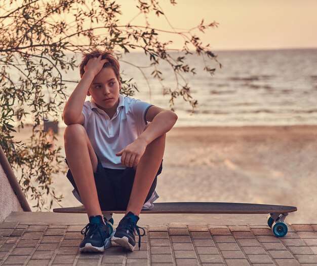 Joven patinador sentado en una patineta contra el fondo de la costa al atardecer.