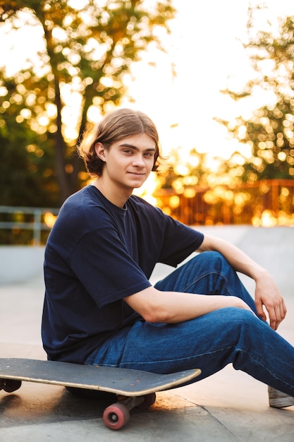 Joven patinador sonriente con camiseta negra y jeans mirando soñadoramente a la cámara pasando tiempo con patineta en el moderno parque de patinaje