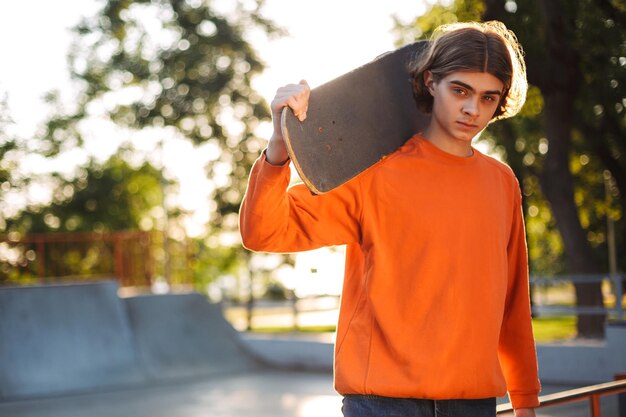 Joven patinador en suéter naranja mirando cuidadosamente a la cámara mientras sostiene el monopatín en el hombro con el parque de patinaje en el fondo