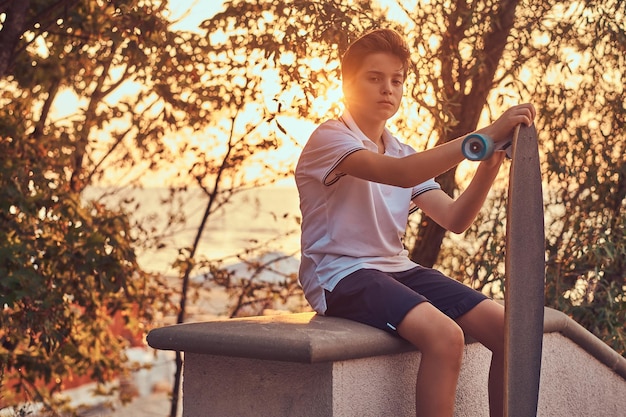 Joven patinador vestido con camiseta y pantalones cortos sentado en la barandilla de piedra al aire libre al atardecer.