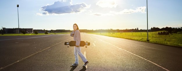 Foto gratuita joven patinadora adolescente patinando en un crucero con una tabla larga y caminando por una carretera vacía de hormigón