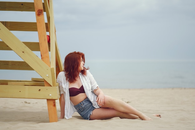 Una joven pelirroja en una camisa blanca y pantalones cortos de jeans sentado en la arena de la playa