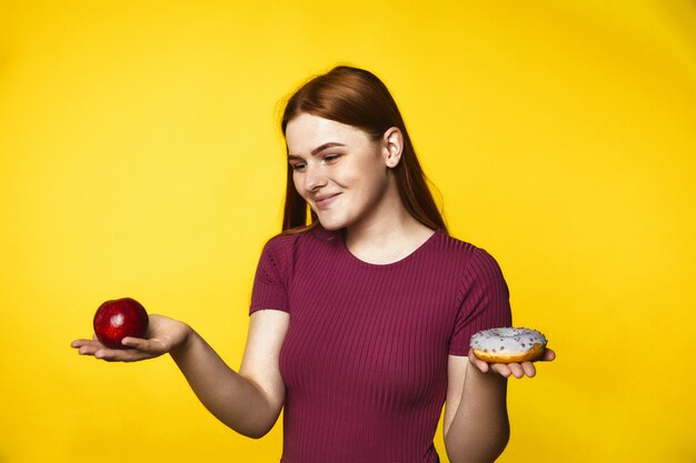 Foto gratuita joven pelirroja elige entre una manzana y una rosquilla