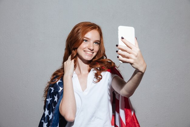 Joven pelirroja feliz con bandera de Estados Unidos hacer selfie