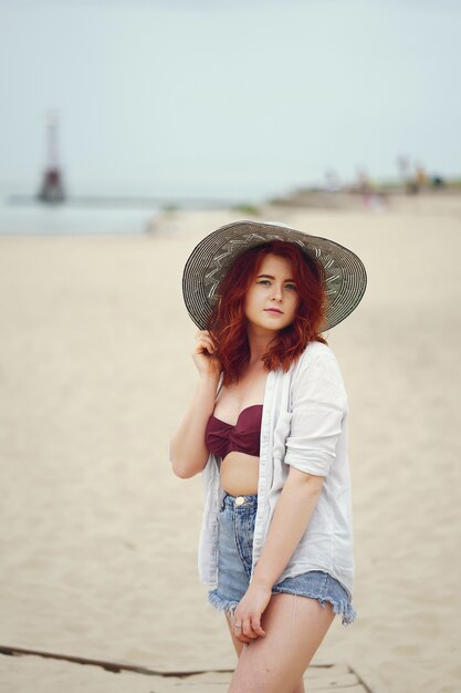 joven pelirroja en un gran sombrero redondo y camisa blanca de pie en la arena de la playa