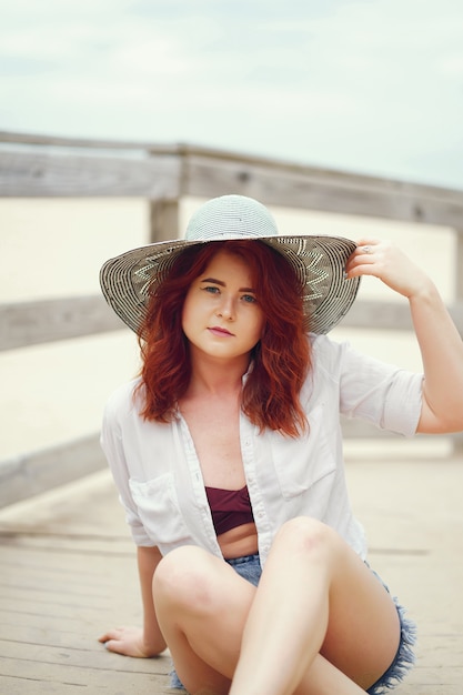 Una joven pelirroja en un gran sombrero redondo sentado en la arena de la playa