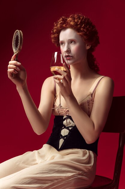 Joven pelirroja medieval como duquesa en corsé negro y ropa de dormir sentada en el espacio rojo con un espejo y una copa de vino