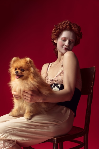 Foto gratuita joven pelirroja medieval como una duquesa en corsé negro y ropa de dormir sentada en una silla en el espacio rojo con un perrito o un perro