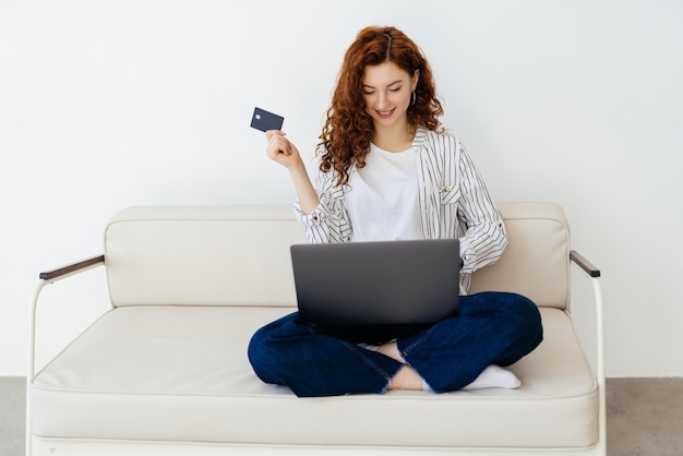 Joven pelirroja relajándose en un sofá en casa con su computadora portátil en tiendas en línea con la información de lectura de su tarjeta de crédito